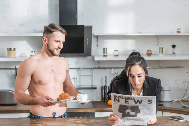 handsome shirtless man holding saucer with croissant and cup of coffee near brunette woman reading business newspaper  - Фото, зображення