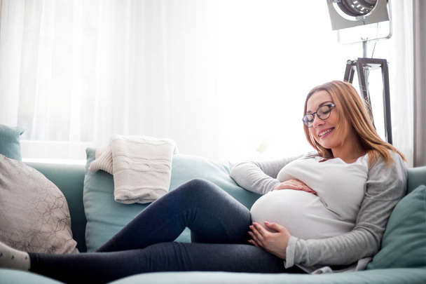 Young pregnant woman sitting on couch in living room - Fotoğraf, Görsel