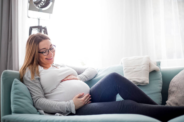 Молодая беременная женщина сидит на диване в гостиной - Фото, изображение