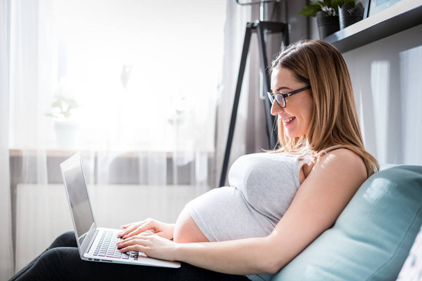 Mulher grávida sorrindo usando laptop, sentado no sofá em casa
 - Foto, Imagem
