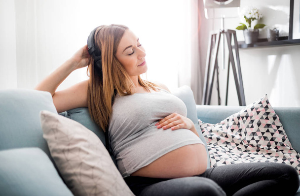 Relaxed pregnant woman sitting on sofa at home and listening music in headphones - Zdjęcie, obraz