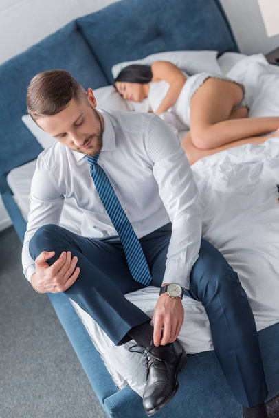 selective focus of bearded man in suit touching shoelaces near sleeping woman  - 写真・画像