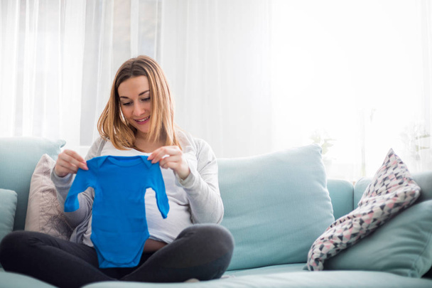 Pregnant woman with baby clothes on her belly, mom expecting child - Zdjęcie, obraz