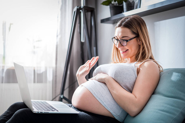 Pregnant woman video chatting with family on laptop waving hand to screen - Valokuva, kuva