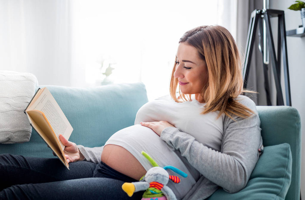 Pregnant woman relaxing on sofa reading book looking at belly - Fotó, kép