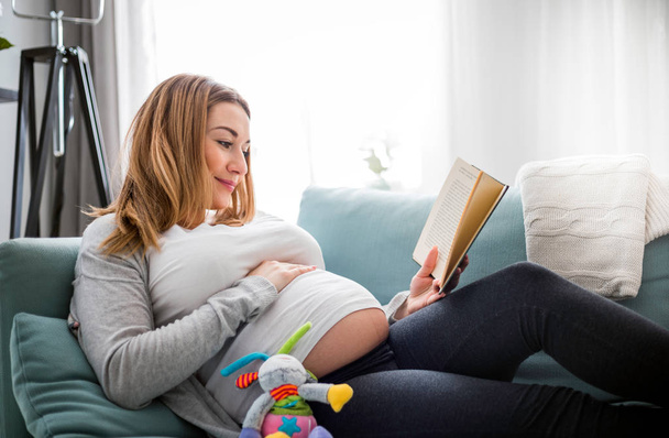 Pregnant woman relaxing on sofa reading book looking at belly - Foto, Imagen