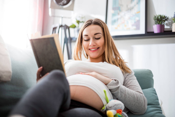 Pregnant woman relaxing on sofa reading book looking at belly - Fotoğraf, Görsel