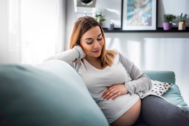 Pregnant woman relaxing on sofa at home in sunny living room - Photo, image