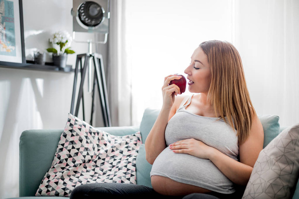 Pregnant woman eating apple and resting sitting on sofa at home - Fotografie, Obrázek