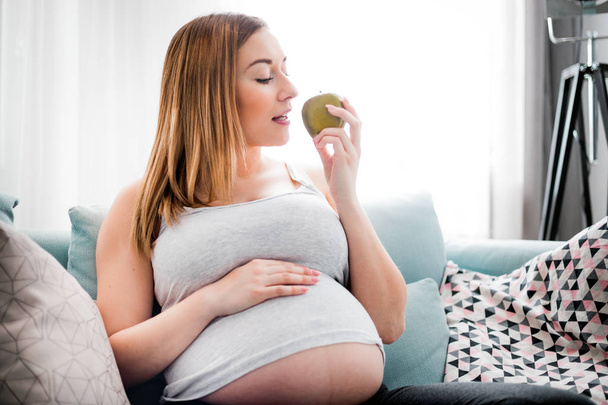 Voeding en voeding tijdens de zwangerschap, zwangere vrouw eten fruit zittend op sofa thuis - Foto, afbeelding