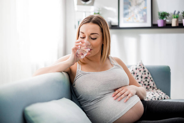 Health during pregnancy, Pregnant woman drinking water sitting on sofa - Fotó, kép