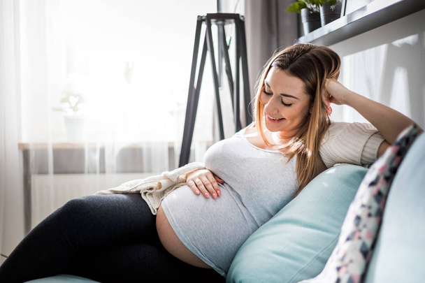 Beautiful pregnant young woman touching her belly sitting on sofa at home - Foto, afbeelding