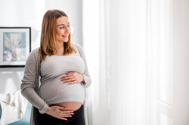 Beautiful pregnant woman touching her belly standing by the window at home - Φωτογραφία, εικόνα