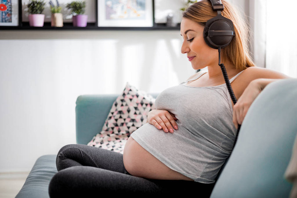 Beautiful pregnant woman sitting on sofa at home and listening music in headphones - Фото, зображення
