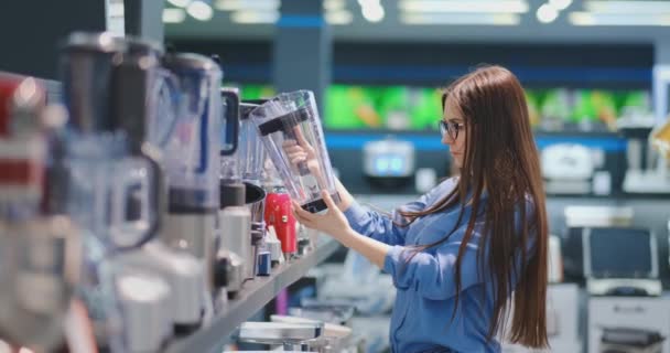 In the appliances store, a brunette woman in a shirt chooses a blender for shopping by viewing and holding the device in her hands. - Filmmaterial, Video
