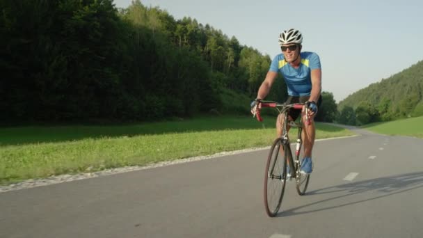 激しい山岳レース中に彼のクールな道路の自転車のペダルを踏んで運動若い男性。日当たりの良い山のプロロードサイクリストトレーニング。夏の自然の中で絵のように美しい自転車に乗る。スローモーション映像 - 映像、動画