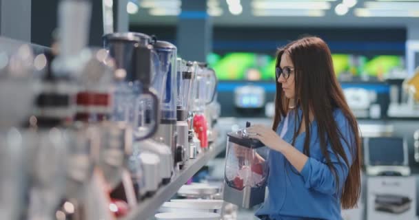 Young beautiful woman in the appliances store is choosing for its kitchen a blender looking and holding in her hands various models. - Imágenes, Vídeo