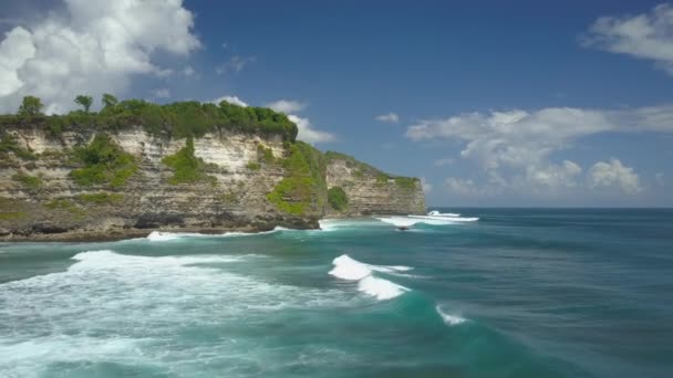 Havadan: Okyanus suyunun köpürttüğü devasa kayalık kayalıklara doğru uçmak. Endonezya'da güneşli bir yaz gününde okyanus suyu kapırıyla yıkanan sakin kayalık sahil şeridinin panoramik manzarası - Video, Çekim