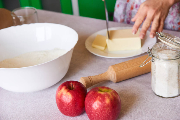 Žena řezaného másla na bílém plechu pro domácí koláč na bílé desky s jablky, válcovým kolíkem a miskou mouky - Fotografie, Obrázek