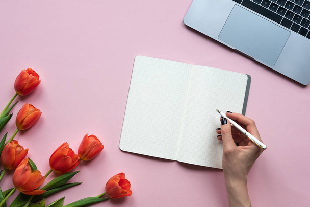 Fechar-se de escrita de mão feminina em um caderno. Fundo rosa com laptop e tulipas. Espaço de cópia
 - Foto, Imagem