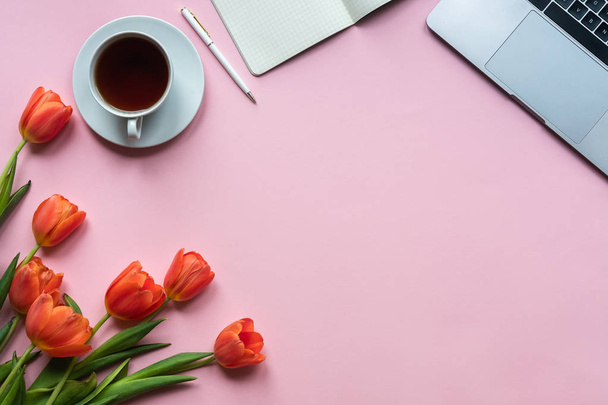 Red tulips with cup of tea, laptop and notebook on pink background. Copy space - Photo, Image