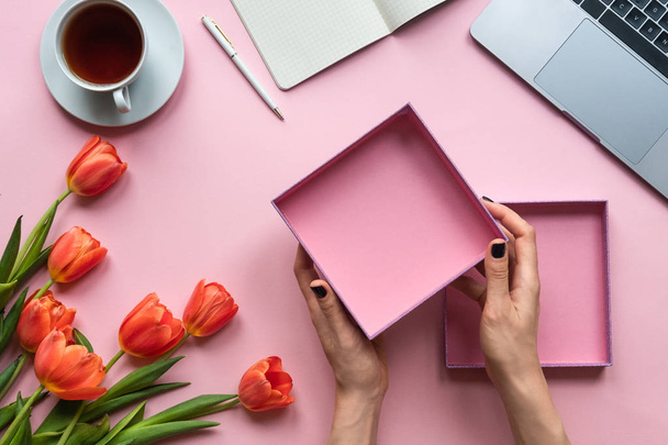 Γυναικεία χέρια που κρατούν ανοιχτό κενό κουτί σε ροζ φόντο. Φόντο με φλιτζάνι τσάι, φορητό υπολογιστή και λουλούδια - Φωτογραφία, εικόνα