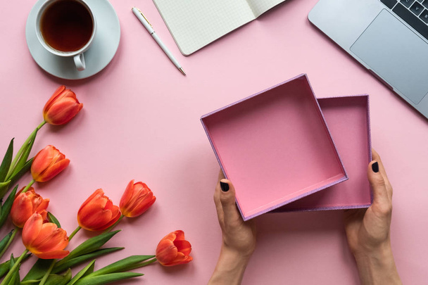Жіночі руки тримають відкриту порожню коробку на рожевому тлі. Фон з кавою, ноутбуком та квітами
 - Фото, зображення