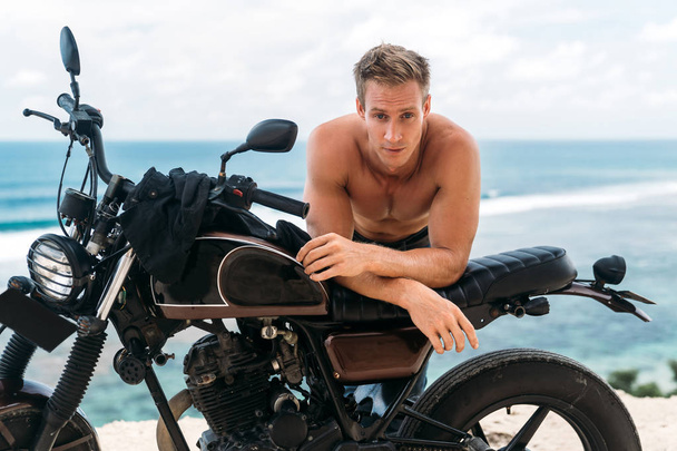 Portrait of sexy athletic man with naked torso on custom motorbike with ocean at background - Photo, Image