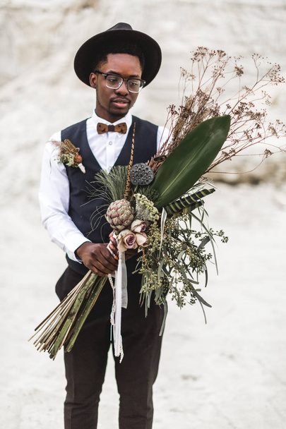 Black Afro American young man standing on the backround of beautiful sand canyon at sunset and holding stylish rustic boho wedding bouquet. Desert wedding, canyon - Foto, Bild