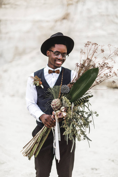 Black Afro American young man standing on the backround of beautiful sand canyon at sunset and holding stylish rustic boho wedding bouquet. Desert wedding, canyon - Фото, зображення