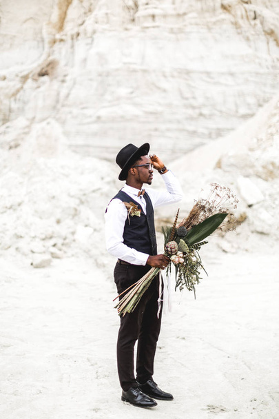 Handsome African man groom in black suit and hat holding wedding bouquet in rustic style and standing in desert canyon against beautiful landscape. - Φωτογραφία, εικόνα