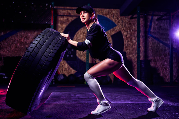 Attractive fit woman athlete working out with a huge tire, turning and flipping in the gym. Cross fit woman exercising with big tire - Foto, immagini