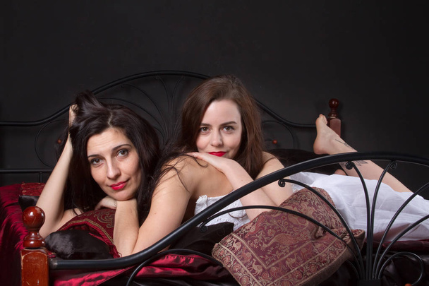 Two young women in vintage lingerie on the spread bed against the black wall, love games, caress, kisses. Added a small grain, imitation of film photography - Zdjęcie, obraz