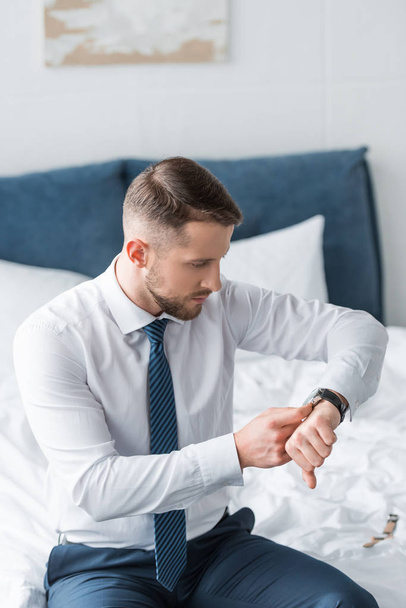 handsome bearded man in formal wear looking at watch while sitting on bed  - Foto, Bild