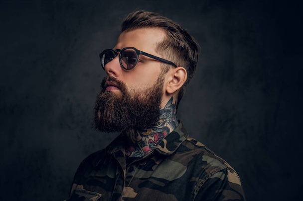 Closeup portrait of a bearded man with a tattoo on his neck in sunglasses wearing a military shirt. Studio photo against dark wall - Photo, Image