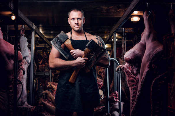 Безрукий татуированный мясник в фартуке позирует с топором на холодильном складе посреди туш мяса
 - Фото, изображение