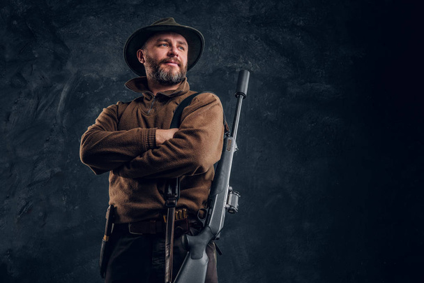 Portrét vousatého lovce s puškou s rukama zkříženýma v ruce. Fotografie ve studiu na tmavém pozadí zdi - Fotografie, Obrázek