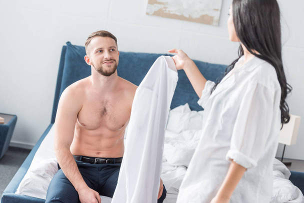 brunette woman holding white shirt near muscular shirtless man sitting on bed - 写真・画像