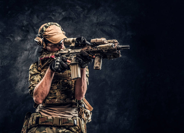 La unidad de élite, soldado de las fuerzas especiales en uniforme de camuflaje que sostiene un rifle de asalto con una mira láser y apunta al objetivo. Foto del estudio contra una pared oscura
   - Foto, Imagen