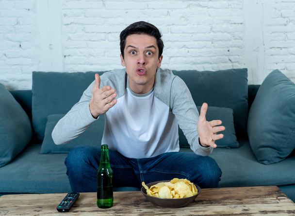 Lifestyle portrait of excited football fan having fun watching soccer or football game on television. Enjoying and celebrating goal and victory drinking beer and eating chips. Sports fans and Goal. - 写真・画像