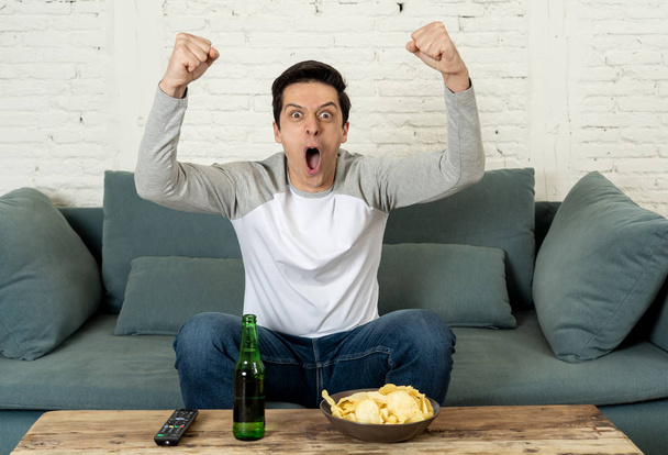 Lifestyle portrait of excited football fan having fun watching soccer or football game on television. Enjoying and celebrating goal and victory drinking beer and eating chips. Sports fans and Goal. - Photo, Image