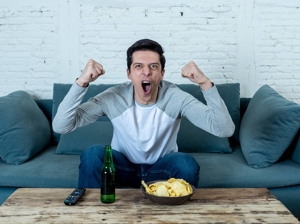 Lifestyle portrait of excited football fan having fun watching soccer or football game on television. Enjoying and celebrating goal and victory drinking beer and eating chips. Sports fans and Goal. - Foto, Imagen
