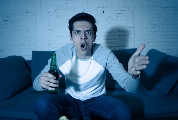 Young man drinking beer at home watching soccer football match or sports game in television at night. Screaming and celebrating goal or victory. In celebration, sports and fans concept. - Фото, зображення