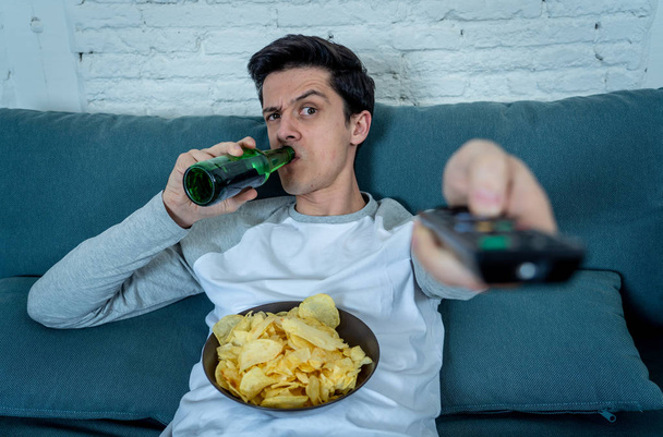 Lifestyle portrait of young bored man on couch with remote control zapping for movie or live sport. Looking disinterested drinking beer. Sedentary and mass social media or Television addiction. - Valokuva, kuva