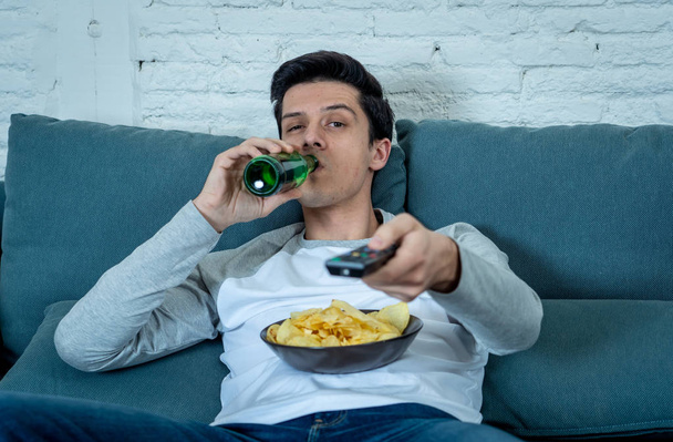 Lifestyle portrait of young bored man on couch with remote control zapping for movie or live sport. Looking disinterested drinking beer. Sedentary and mass social media or Television addiction. - Photo, Image