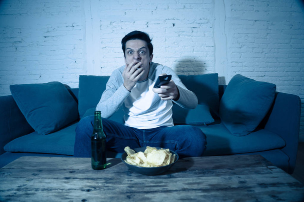 Lifestyle portrait of young scared man watching horror movie or thriller on TV. Holding remote control shocked making frightened gestures. In people, mass media, television and entertainment concept. - Foto, Imagem