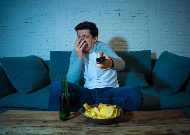 Lifestyle portrait of young scared man watching horror movie or thriller on TV. Holding remote control shocked making frightened gestures. In people, mass media, television and entertainment concept. - Photo, Image