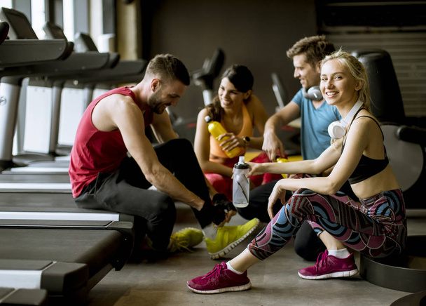 Groep jonge mensen in sportkleding praten en lachen samen terwijl zittend op de vloer van een sportschool na een training - Foto, afbeelding