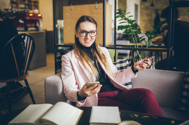 theme modern profession female blogger. Caucasian woman with glasses and jacket sitting inside coffee shop behind wooden table with notebook, laptop and cup coffee. Girl emotion dream, think writer - Photo, Image