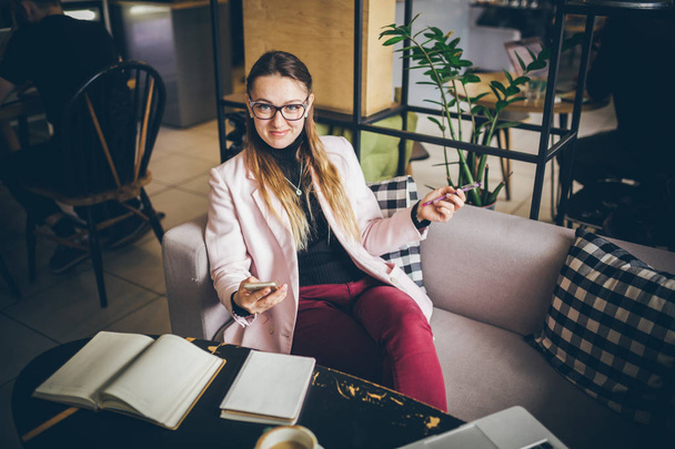 theme modern profession female blogger. Caucasian woman with glasses and jacket sitting inside coffee shop behind wooden table with notebook, laptop and cup coffee. Girl emotion dream, think writer - Фото, зображення